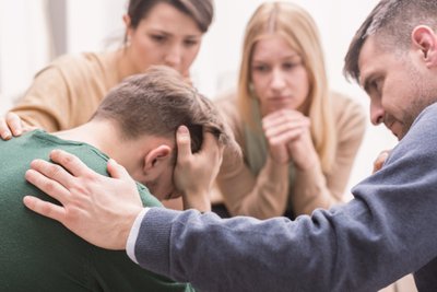 Close-up of a devastated young man holding his head in his hands and friends supporting him during group therapy